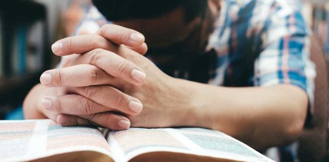 Man Praying Hands Clasped On Bible iStock-938108512