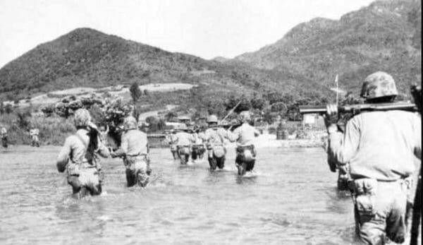 Members of the 5th Marines slog through a flooded area of Korea on the way back from a day of training. Through thigh-high water carrying weapons on their backs, walking toward a hill in the distance.